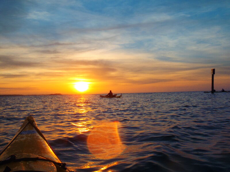 The image shows a serene ocean scene at sunset. A person is kayaking in the distance, silhouetted against the bright sun. The sky is a mix of orange, yellow, and blue hues, reflecting on the water's surface. The front of a yellow kayak is visible in the foreground, adding depth to the composition.